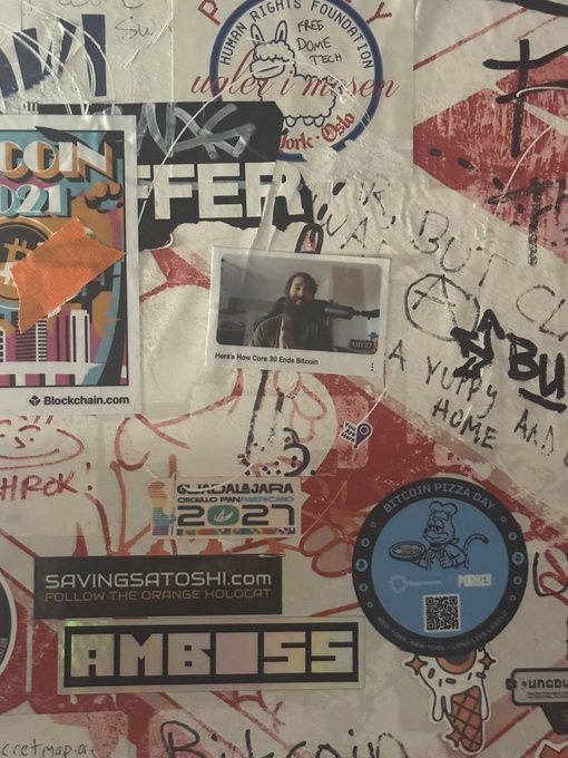 A wall covered in graffiti and overlapping crypto-themed stickers—including "Bitcoin Pizza Day," "Blockchain.com," "AMBOSS," "Satoshi Free Dome," and "Bitcoin 2021 Miami"—with a Polaroid labeled "Here's How Core 30 Ends Bitcoin" placed centrally, embodying the spirit of decentralized expression by treating public sticker art as graffiti permanently etched onto the blockchain.