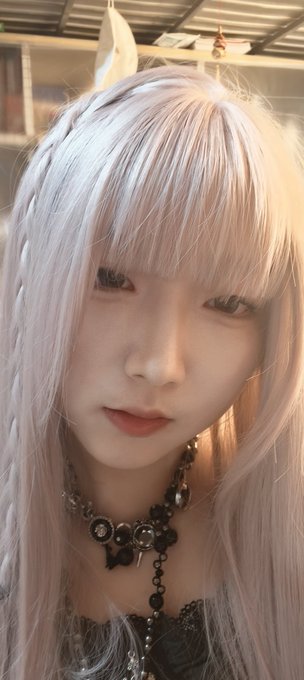A selfie-style portrait of a young woman with long light pink/lavender hair wearing a gothic-style black necklace.