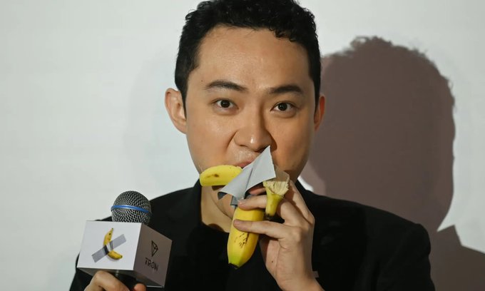 A man with short black hair, wearing a dark shirt, is shown eating a banana that has been modified to display the TRON logo, while holding a microphone branded with the same TRON insignia. The image illustrates the caption's narrative about Justin Sun, the founder of TRON, and his controversial tactics involving crypto exchanges like HTX, Binance, and Coinbase.