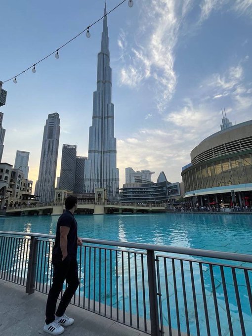 A person stands by a railing overlooking a body of water with the Burj Khalifa towering in the background in Dubai. String lights hang overhead, and a building labeled "EMAAR" is visible among the skyline.