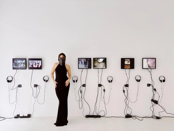 A woman in a black dress and face mask stands in a white gallery space, posing beside an art installation featuring six wall-mounted screens with headphones and tangled cords hanging beneath them.