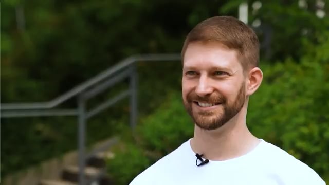 A portrait of a smiling man with short reddish-brown hair and a beard, wearing a white t-shirt with a small microphone clipped to it, standing outdoors with a blurred green background.