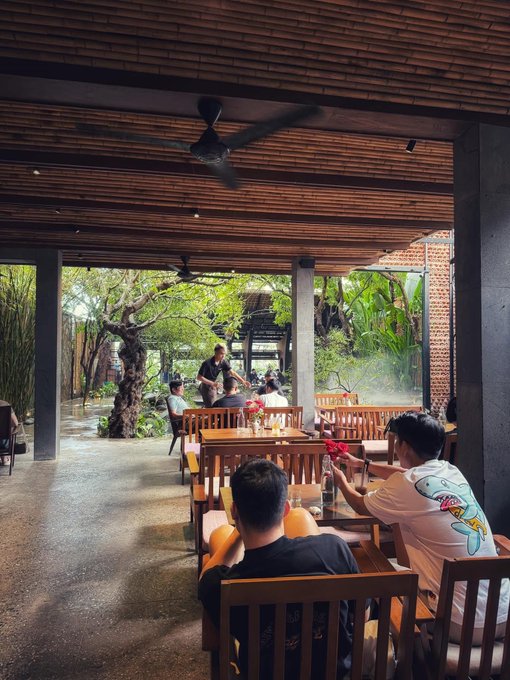 A serene open-air café or restaurant with a wooden slatted ceiling and ceiling fans, where patrons sit at wooden tables surrounded by lush green trees and plants on a bright day.