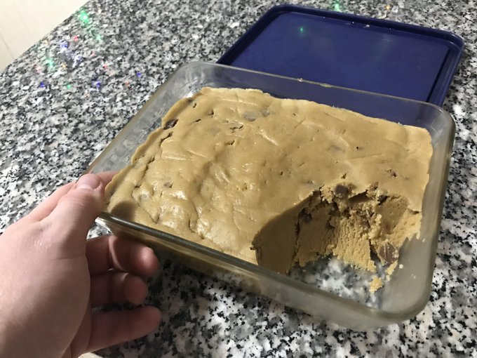 The image features a person holding a glass dish filled with cookie dough or cake batter, which appears to be a chocolate chip cookie. The person's hand is reaching out towards the dish, possibly about to take a bite of the dessert. The background shows a countertop made of granite tiles, providing a contrast between the softness of the baked goods and the hard surface beneath them.