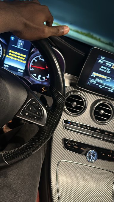 A person grips the steering wheel of a Mercedes-Benz, with the digital dashboard displaying an active radio station.