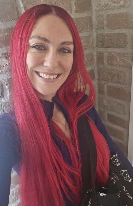 A smiling woman with long bright red hair wearing a dark jacket poses in front of a brick wall background.