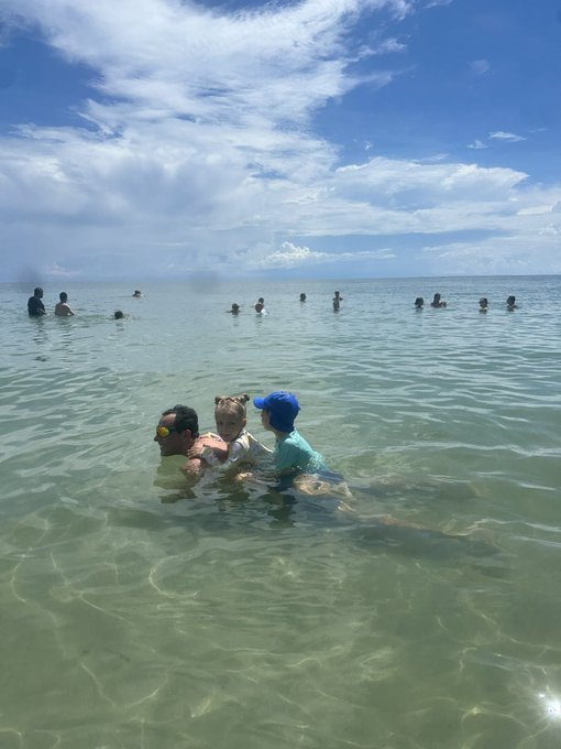 A family enjoys crystal-clear turquoise waters on a sunny Florida beach day, with several other swimmers visible in the distance.

The image supports the post's NYC-to-Miami relocation theme by showcasing the quintessential Miami lifestyle—relaxed beach time, warm weather, and waterfront leisure—that contrasts with the faster-paced NYC culture mentioned in the cons.