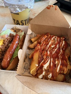 The image shows a box of food, including a hot dog with ketchup and mustard as well as some fries covered in ketchup. The food items are placed next to each other inside a cardboard container or box. There is also a drink present in the scene, which appears to be a cup. The overall setting suggests that this meal could be enjoyed during a lunch break or while watching a movie at home.