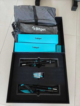 An opened Bitget gift box containing a gray branded bag, two teal Bitget-branded boxes, and what appears to be a tripod in foam packaging.