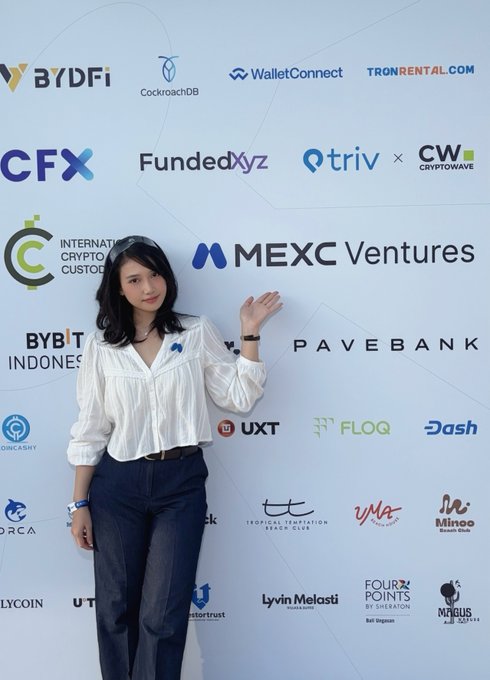 A woman wearing a white shirt and sunglasses on her head standing in front of a sponsor wall featuring crypto and fintech logos including BYDFi, CockroachDB, WalletConnect, MEXC Ventures, Pavebank, and others.