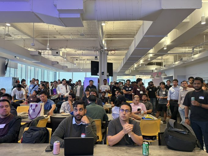 A packed room of attendees at the Cafe Compute NYC event, with many standing in the back despite the tornado warning mentioned in the post. Visible details include a laptop on the foreground table, a green beverage can, and name tags on attendees, illustrating the high turnout and enthusiasm for AI/tech networking described in the caption.