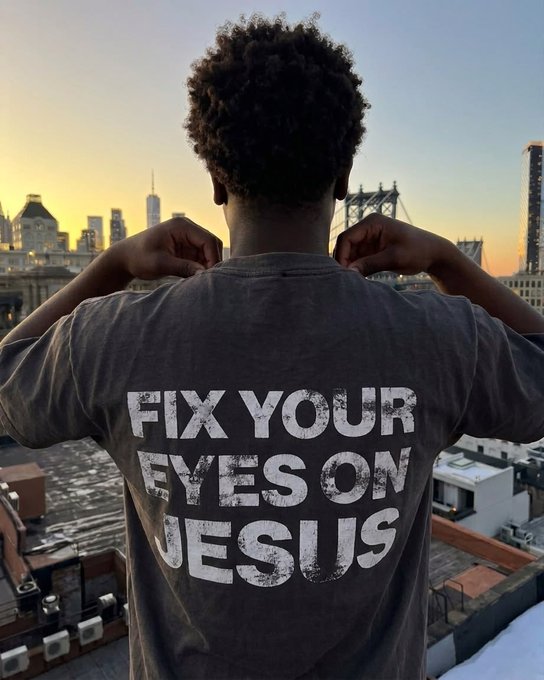 A person viewed from behind wears a grey shirt reading 'FIX YOUR EYES ON JESUS' while looking out over a city skyline at sunset.