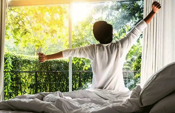 A person sits on a bed with arms outstretched, stretching toward a large window with a bright, sunlit garden view.