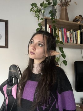 A young woman with long dark hair sits in front of a bookshelf and plants, wearing a purple and black striped sweater.