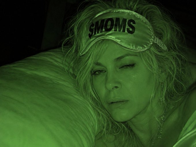 A green-tinted close-up photo of a woman wearing a sleep mask that reads '$MOMS', lying on a pillow.