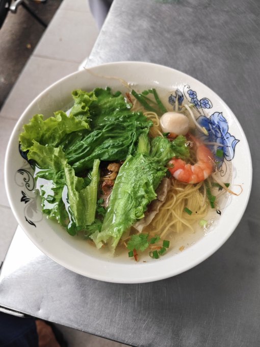 A bowl of noodle soup containing shrimp, lettuce, herbs, and other ingredients served on a metal table.