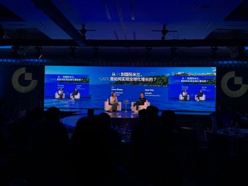 A wide shot of a conference or event stage with blue lighting, featuring two speakers seated at a table in front of large screens displaying event branding.