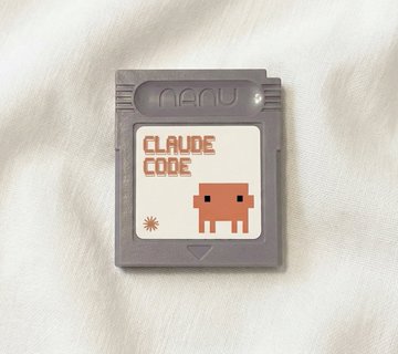A photo of a gray Nintendo Game Boy cartridge with an orange label reading 'CLAUDE CODE' featuring a small pixel-art robot character, resting on a white fabric surface.