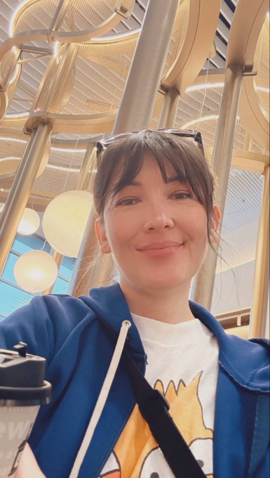 A selfie of a smiling woman wearing a blue hoodie and white t-shirt, with sunglasses on her head, in front of an ornate ceiling with golden arches and round light fixtures.