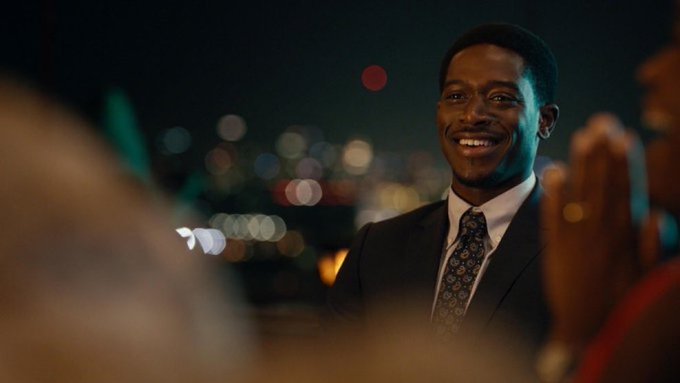 A smiling Black man in a suit and patterned tie at a nighttime outdoor event, with blurred city lights in the background.