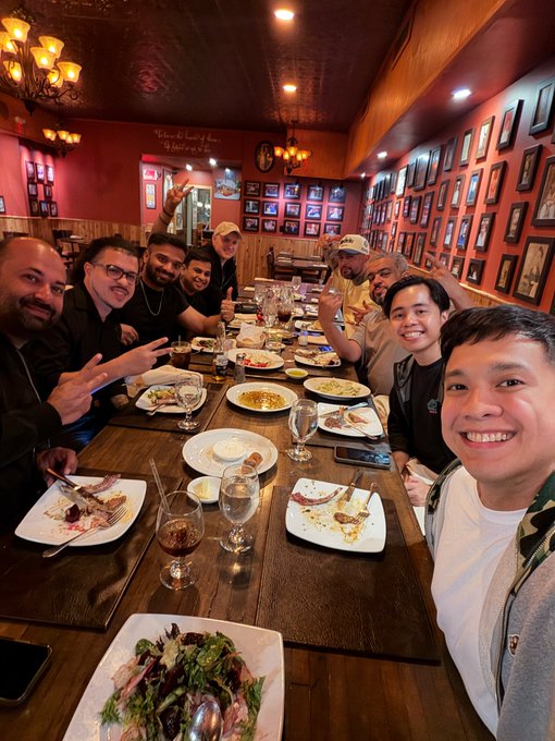 A group of friends poses cheerfully at a long dining table covered with empty plates and wine glasses, with a fresh salad in the foreground and warmly lit restaurant decor featuring framed pictures on the walls in the background, capturing the joyful dinner gathering highlighted in the post at Destefanos Steakhouse.