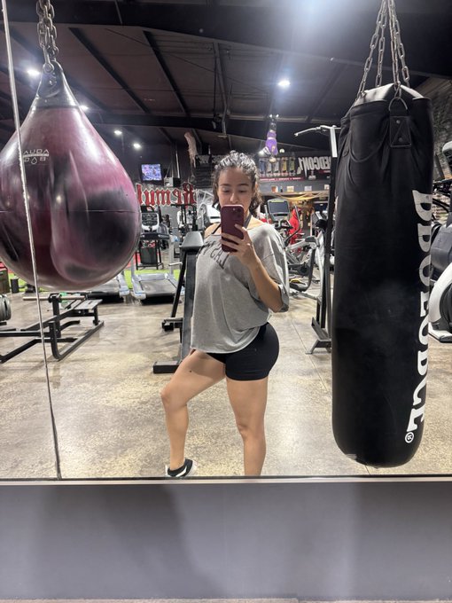 A woman taking a mirror selfie in a gym, standing between a purple water-filled punching bag and a black heavy bag, wearing a gray sweatshirt and black shorts.