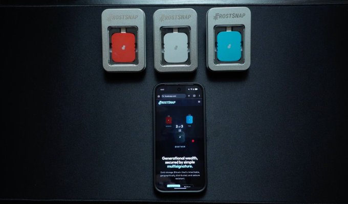 Three Frostsnap hardware signing devices—red, silver, and teal—are arranged above a smartphone displaying the Frostsnap app's 2-of-3 multisignature setup screen, illustrating the multi-device approach highlighted in the review of the cold storage wallet.