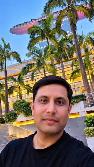 A man with short dark hair and a black t-shirt takes a selfie in front of a modern building with curved architecture and palm trees at dusk.