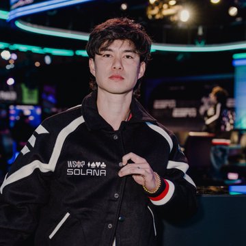 MINHxDYNASTY stands at a crypto trading desk wearing a black “SOLANA” jacket, pictured in connection with his journey from losing $50K and getting scammed to turning a $1K recovery into a $500K portfolio.