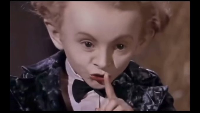 A close-up of a young boy with distinctive facial features wearing a tuxedo, holding a finger to his lips in a 'shh' gesture.