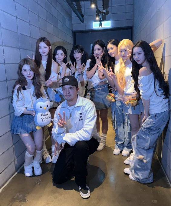 A group of eight young women and one man pose for a photo in a narrow, tiled hallway, with the man crouching in the center and one woman holding a plush penguin toy labeled 'KING PENGUIN'.