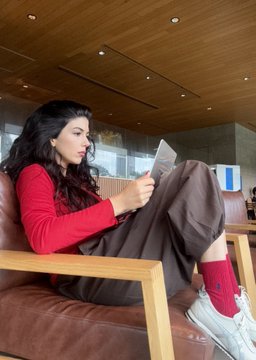 A woman with long dark hair sits on a brown couch reading a booklet, wearing a red top and brown pants with red socks.