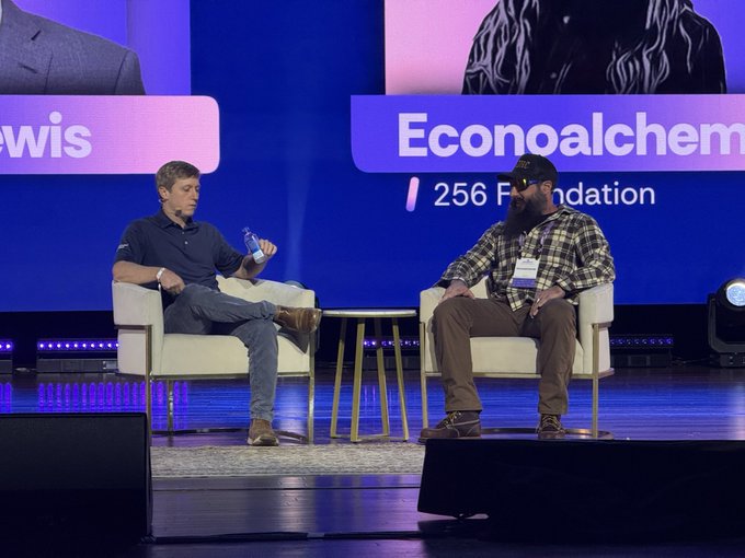 Two men are seated in white chairs on stage during a panel discussion, with one wearing a navy polo holding a water bottle and the other in a plaid shirt and cap. The backdrop displays partial text reading "Econoalchemy" and "256 Fo[undation]," corresponding to the 256 Foundation's presence at the event.