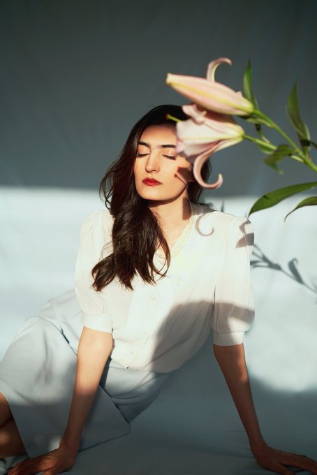 The image features a woman sitting cross-legged with her legs spread out, wearing a white shirt and skirt. She has long hair that falls over her shoulders. A pink flower is positioned above her head, creating an interesting visual effect as she looks at it. The background of the photo appears to be blue or gray, which complements the color of the flower. There are no texts present in the image, but there is a caption that reads "GN Art Lovers Between Light and Bloom 8/10 available for 0.01 $ETH will appreciate any supports".