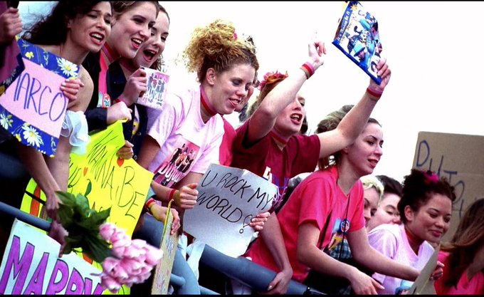An enthusiastic crowd of young women hold up colorful signs with text including 'MARCO', 'BE', 'UNLOCK MY WORLD', and 'MADONNA'.