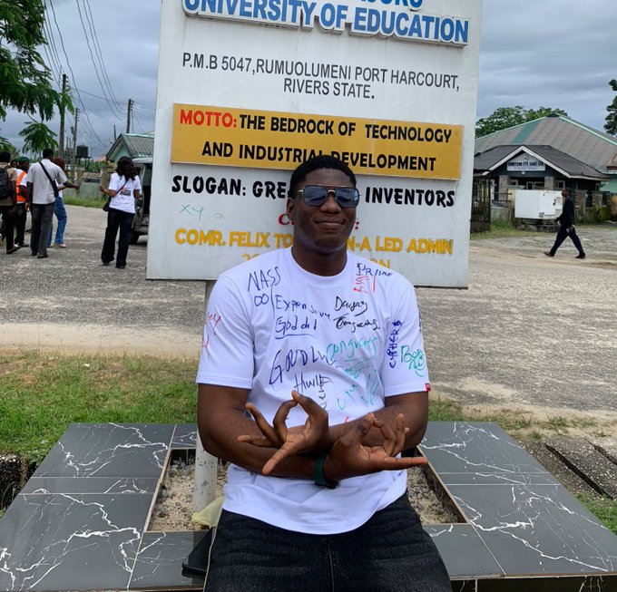 A young man wearing sunglasses and a white t-shirt covered in handwritten messages sits in front of a sign for a University of Education in Rivers State, with the motto 'THE BEDROCK OF TECHNOLOGY AND INDUSTRIAL DEVELOPMENT'.