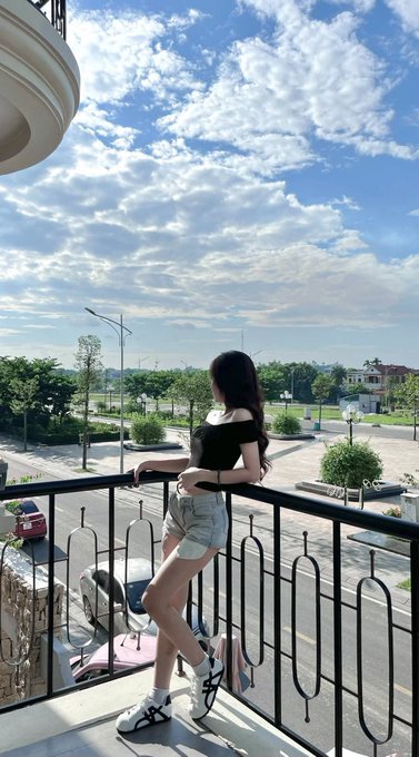 A young woman with long dark hair poses on a balcony overlooking a street, wearing a black top, denim shorts, and chunky black and white sneakers.