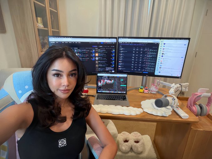 A young woman takes a selfie in front of an elaborate trading setup featuring three monitors displaying crypto charts and trading platforms, a laptop with additional market data, and pink gaming headphones — visually reinforcing the joke that sacrificing a personal life is a hidden "risk" of dedicating oneself to crypto trading.