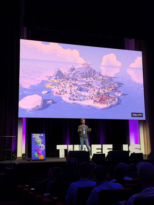 A presenter stands on stage at a "THREEF IS" event showcasing a large screen displaying the colorful, isometric landscape of Pudgy World, with a smaller Pudgy Penguins logo visible on a side display to an audience in a darkened auditorium.
