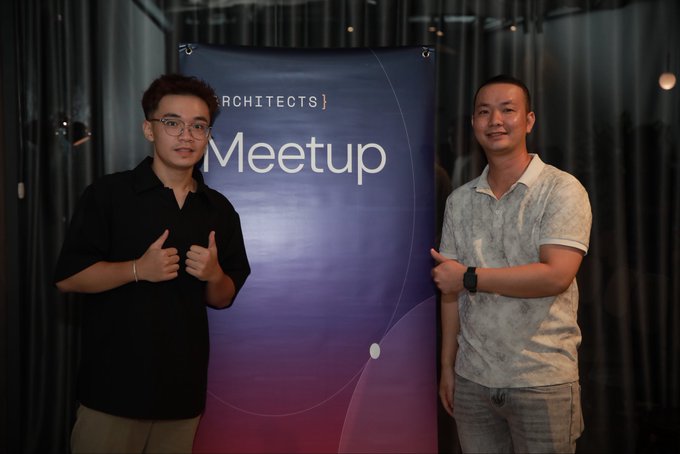 Two men smiling and giving thumbs up in front of a banner that reads 'RCHITECTS Meetup', with the younger man on the left wearing glasses and a black shirt, and the older man on the right wearing a patterned polo shirt.