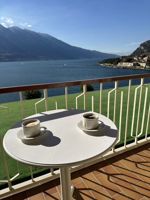 Two espresso cups sit on a small round table on a balcony overlooking a large blue lake surrounded by mountains and a distant village.