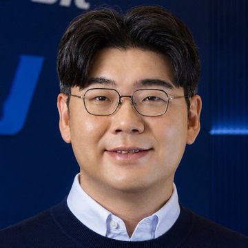 A headshot of a smiling man with glasses wearing a dark sweater over a light blue collared shirt against a blue background.