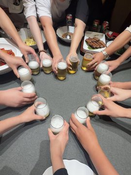 A group of people gathered around a circular table raising their glasses of beer in a toast, with plates of food visible on the table in a social gathering setting.