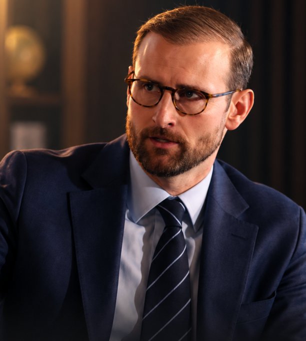 A man in a dark suit, light blue shirt, and navy striped tie wearing tortoiseshell glasses is pictured in what appears to be an office setting with a globe visible in the background, corresponding to the post about White House Executive Director Patrick Witt urging lawmakers to act on the Clarity Act.