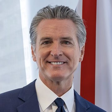 California Governor Gavin Newsom smiles at the camera, dressed in a dark suit and blue tie, with the American flag and what appears to be the California state flag visible behind him. The image accompanies news of his new legislation banning state officials from issuing memecoins and establishing guidelines to protect crypto scam victims.