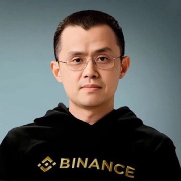 A man wearing glasses and a black 'BINANCE' hoodie poses for a portrait against a gray background.