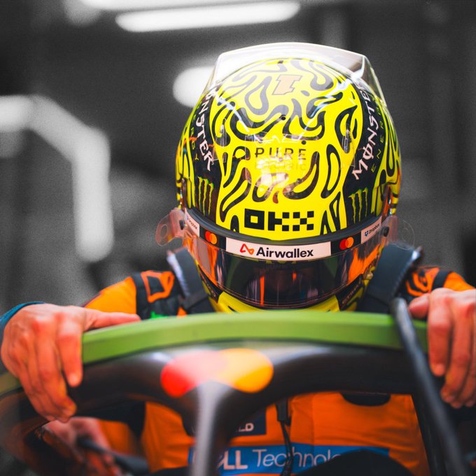 A close-up shows a racing driver gripping a green steering wheel inside a car, wearing a bright yellow helmet covered in black flame-like graphics and sponsor logos. Visible branding includes “Airwallex” on the helmet visor strip and “AII Technologies” on the suit, matching the post’s reference to loading up for Singapore.