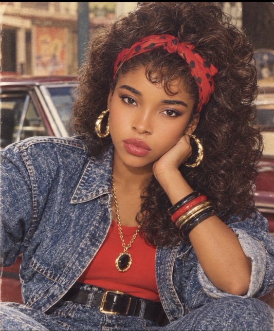 A portrait of a young woman with voluminous curly hair wearing a red polka-dot bandana, denim jacket, red top, gold hoop earrings, and stacked bangles in a retro 80s/90s style.