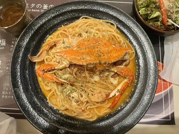 A black bowl containing spaghetti in a creamy orange sauce topped with crab legs and herbs, served alongside a glass of iced drink and a salad bowl on what appears to be a Japanese restaurant tray.