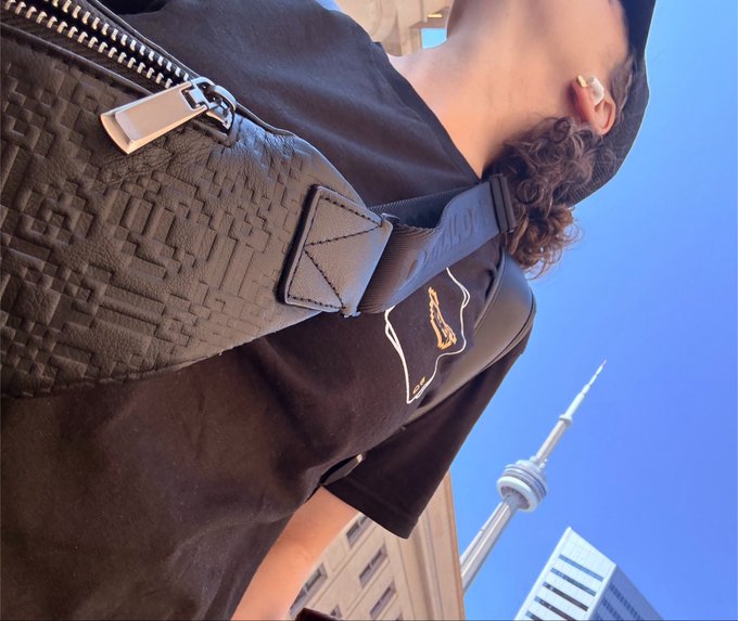 A low-angle selfie showing a person wearing a black t-shirt with a Louis Vuitton bag strap visible, with the CN Tower in the background against a blue sky.